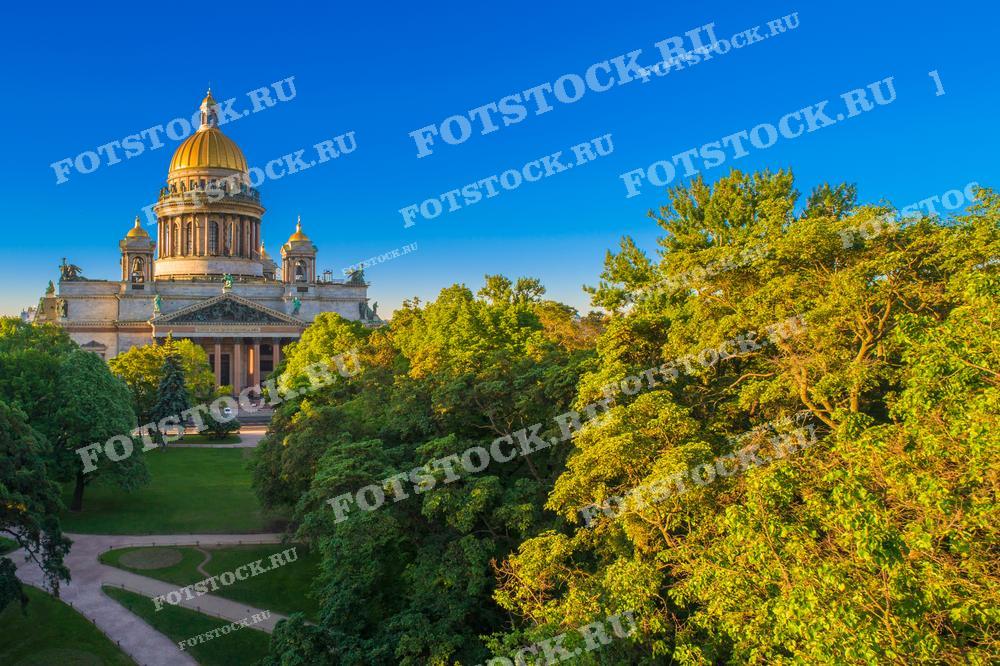 Эта фотография представляет величественный вид на Исаакиевский собор в Санкт-Петербурге, открывающийся через зелёные насаждения парка. Золотой купол собора сверкает на солнце, выделяясь на фоне ясного голубого неба. Такое изображение может быть использовано для туристических гидов, культурно-исторических материалов, а также для декора интерьеров.
